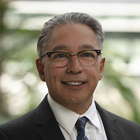 Portrait of professor Héctor González wearing eye glasses, white shirt, dark tie and black jacket.