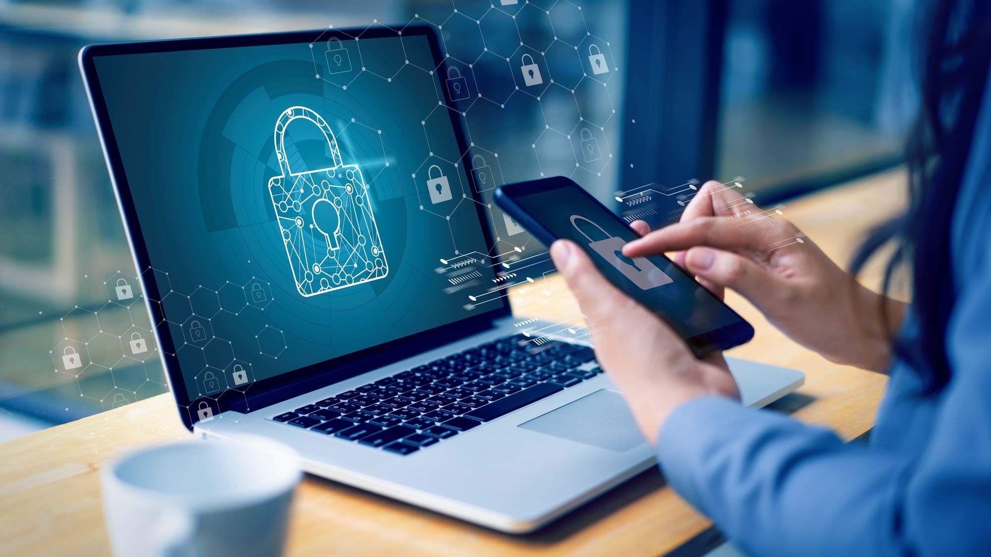 A woman's hands holding a smartphone with a lock symbol on it, in front of a laptop that also has a lock symbol on it.