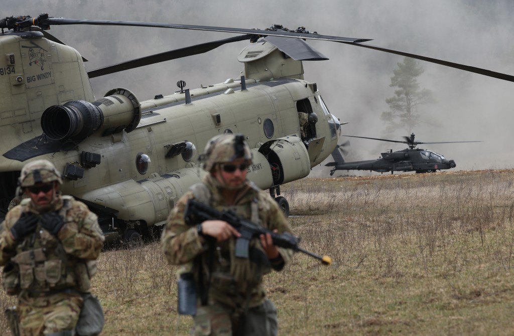 Two US Army soldiers in combat gear in a field with a Chinook helicopter and an Apache helicopter in the background during the Allied Spirit 25 military exercise.