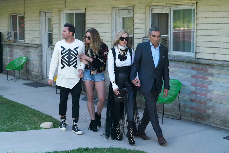 Four people stand in front of a motel. One wears a patterned sweater, another a fur coat with shorts, one in a black and white ensemble, another in a suit