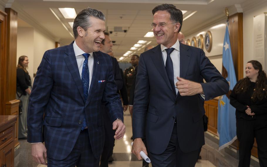 Two men in suits walk side by side down a hallway, smiling.