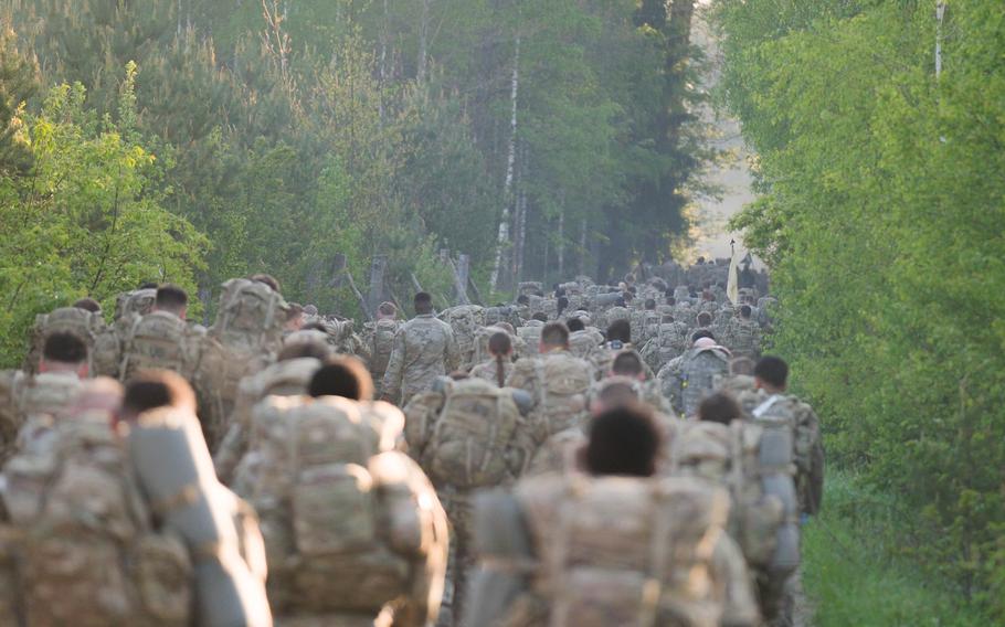 A long line of soldiers in camouflage uniforms with rucksacks marches down a tree-lined road.