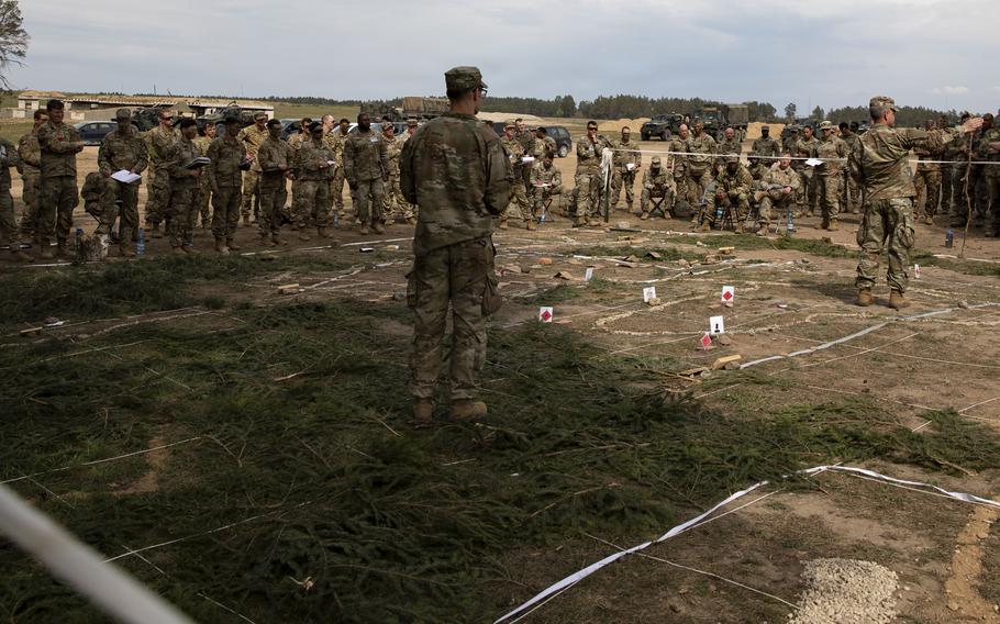 A soldier in camouflage stands with his back to the camera at the front of a large outdoor formation of troops gathered around a terrain model in a grassy field.