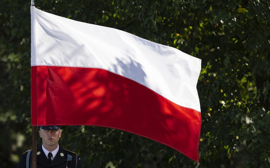 A soldier in dress uniform carries a Polish flag with white and red horizontal stripes, framed by green trees in the background.