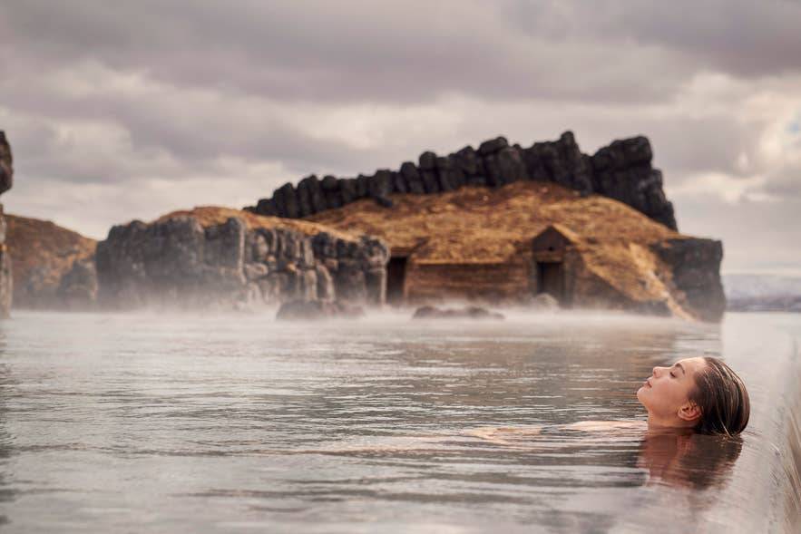 Woman relaxing in the warm waters of Sky Lagoon, Iceland, with dramatic rocky cliffs and turf houses in the background. Woman relaxing in the warm waters of Sky Lagoon, Iceland, with dramatic rocky cliffs and turf houses in the background.