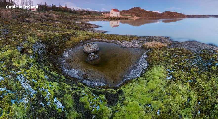 Moss-covered lava rocks and calm waters in East Iceland, with a white house reflected in the still lake at sunset. Moss-covered lava rocks and calm waters in East Iceland, with a white house reflected in the still lake at sunset.