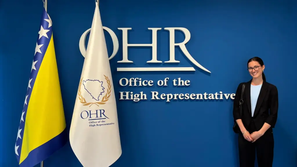Asja Tuco stands near flags and writing on the wall that reads OHR Office of the High Representative
