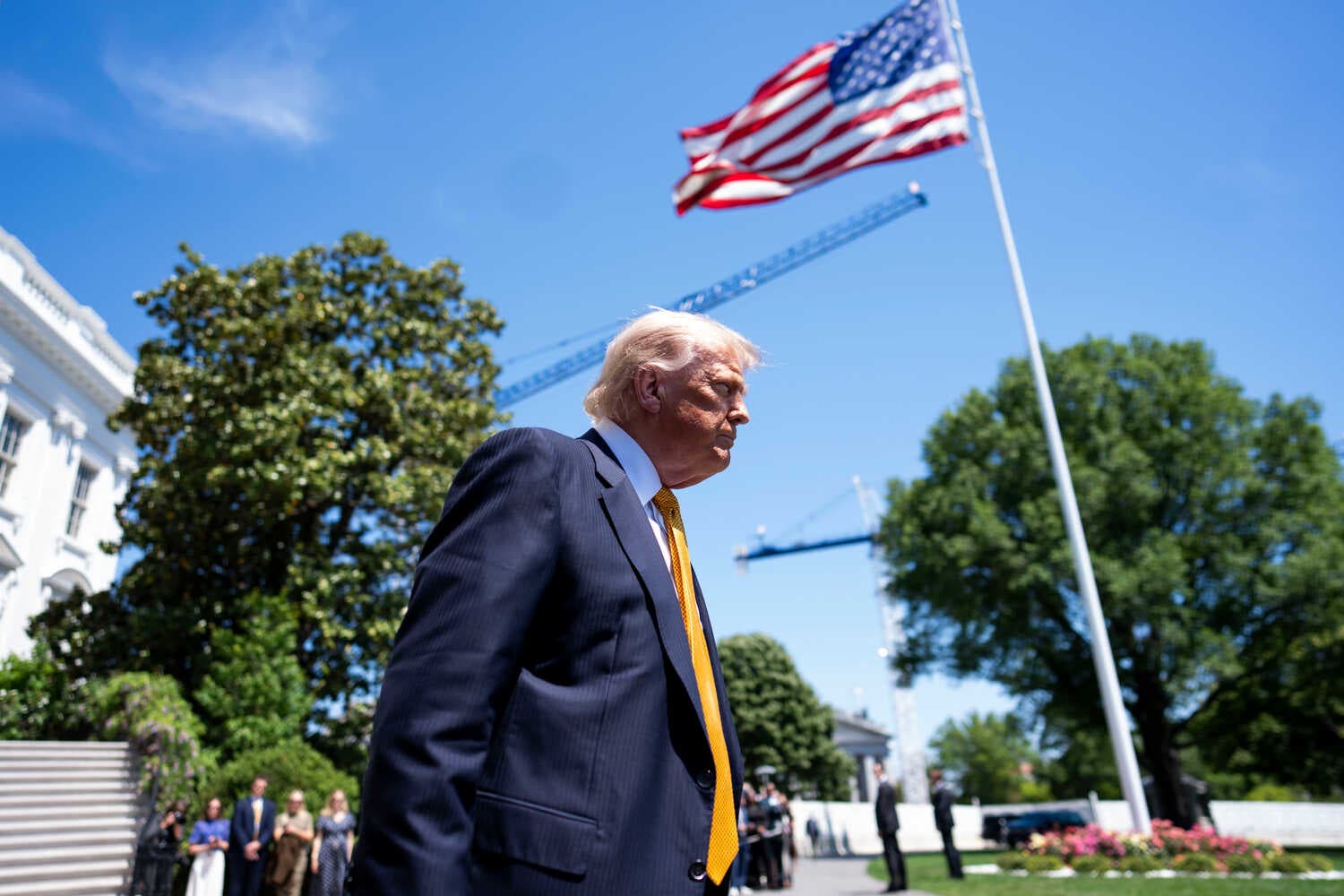 Donald Trumps in a navy blue jacket and gold tie walks underneath an American flag outside the white house. 