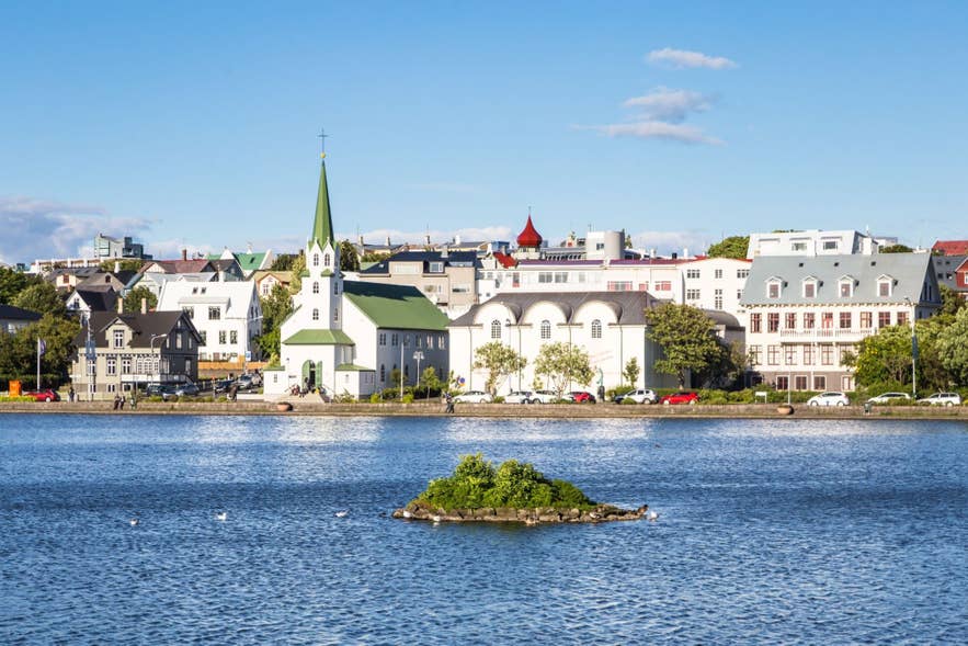 View across Tjornin lake toward downtown Reykjavik, with colorful buildings and Frikirkjan church reflected along the waterfront.