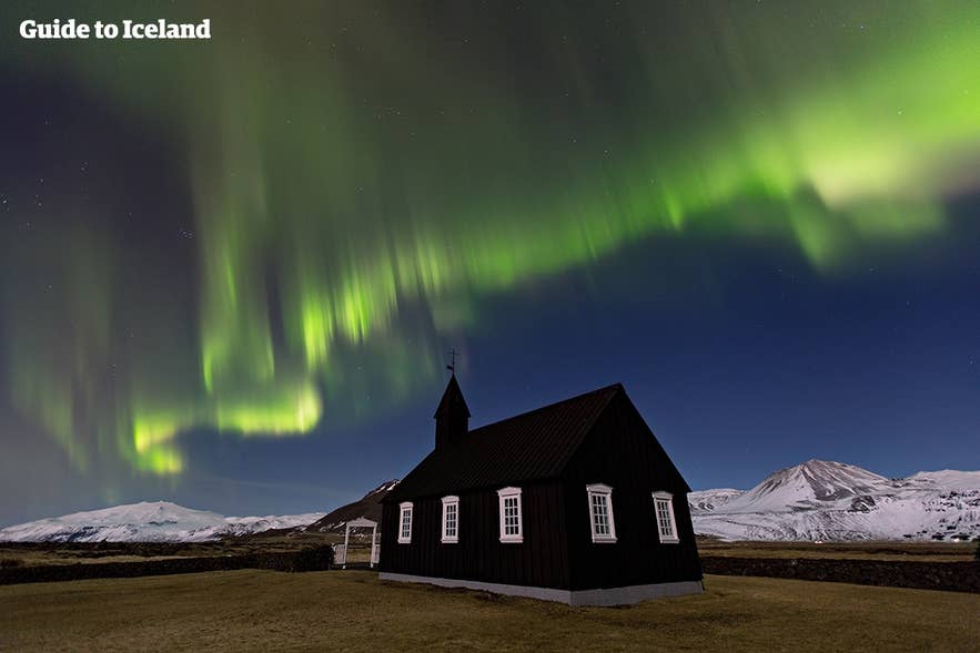 Northern lights over the black church of Budir in Iceland, with snowy mountains and a clear night sky in the background. Northern lights over the black church of Budir in Iceland, with snowy mountains and a clear night sky in the background.