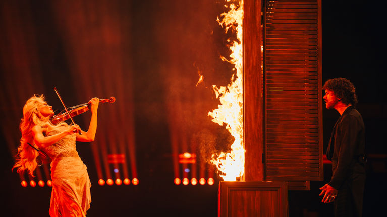 Woman with long blonde hair playing the violin on a dark stage, opposite a man in dark clothing looking at her through a window of flames.