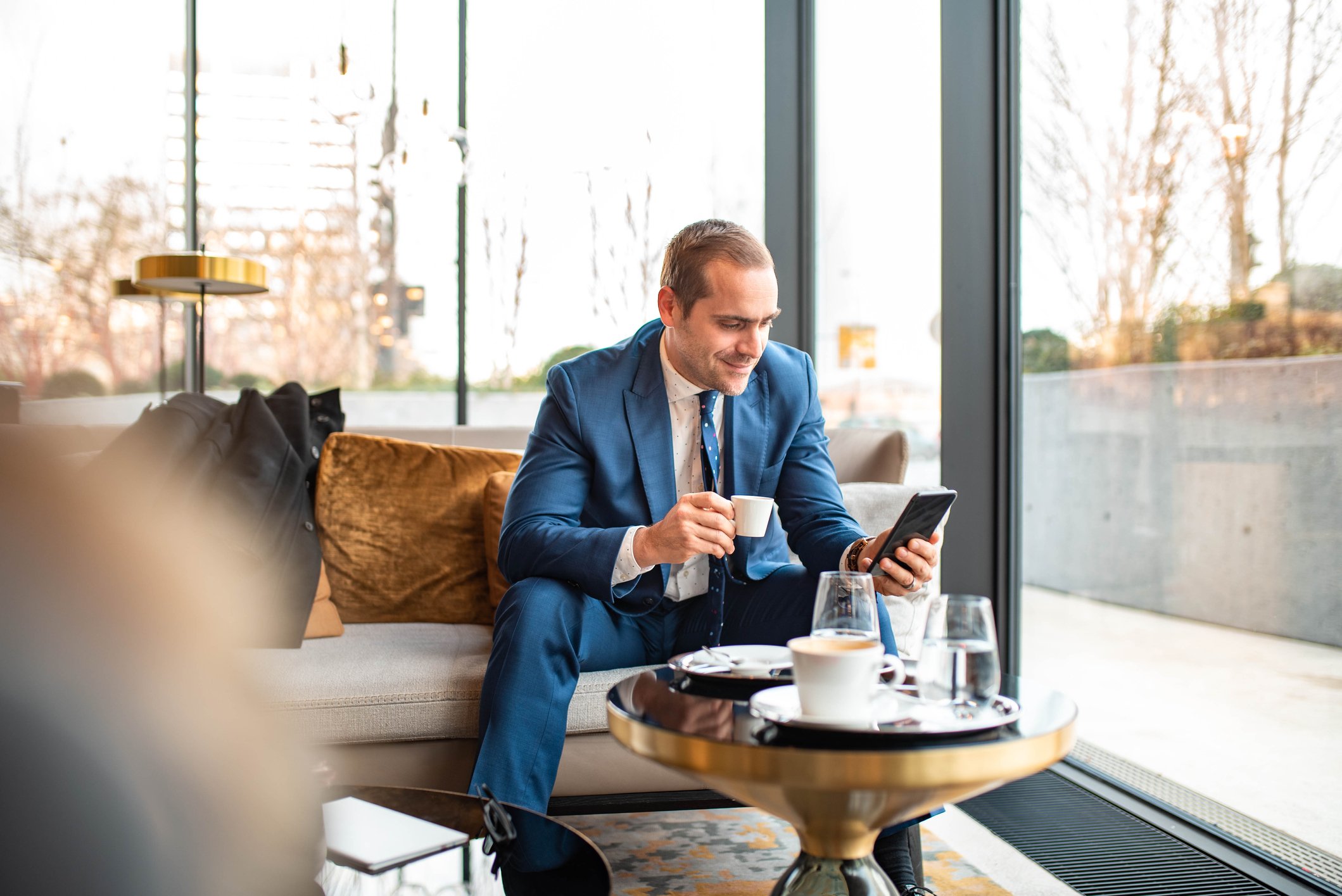An investor sits in a cafe and smiles while looking at a cell phone and sipping from an espresso cup.