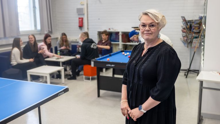Henkilö hymyilee kameralle, taustalla opiskelijoita.