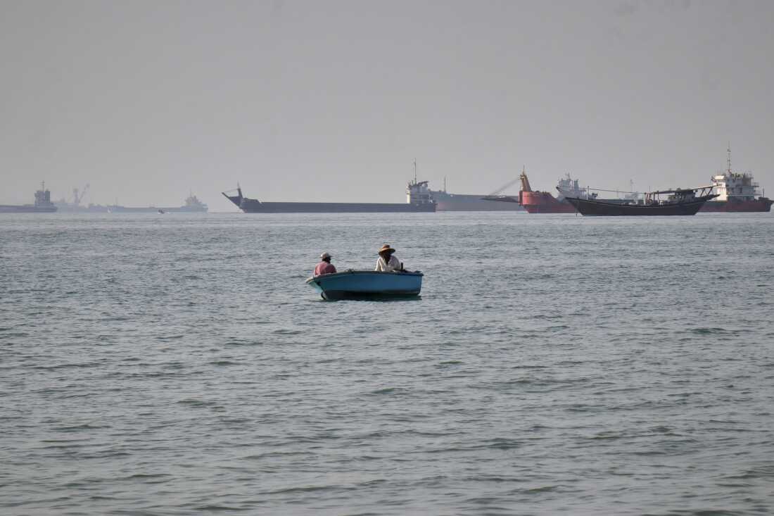 Two men sit in a small boat on the water as a mix of bulk carriers, cargo ships, and service vessels line the horizon in the Strait of Hormuz off Bandar Abbas, Iran, Monday, April 27, 2026.