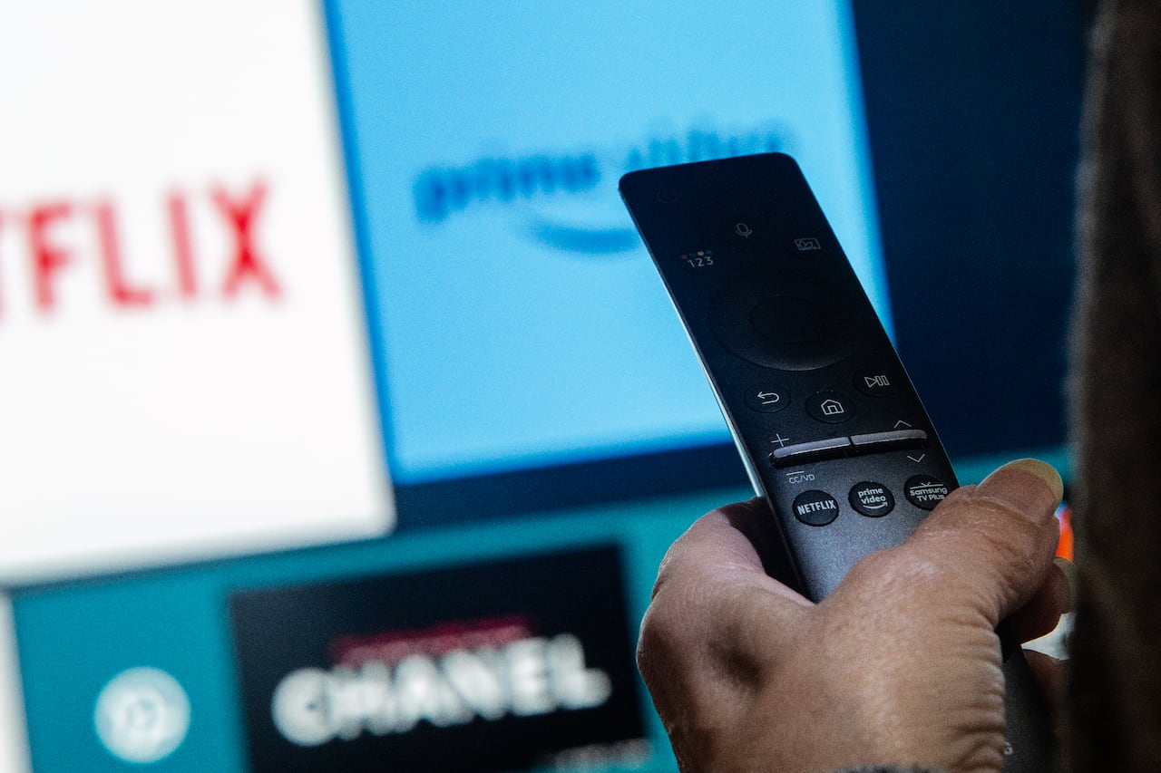 A hand holds a remote control as a TV screen in the background displays streaming services.