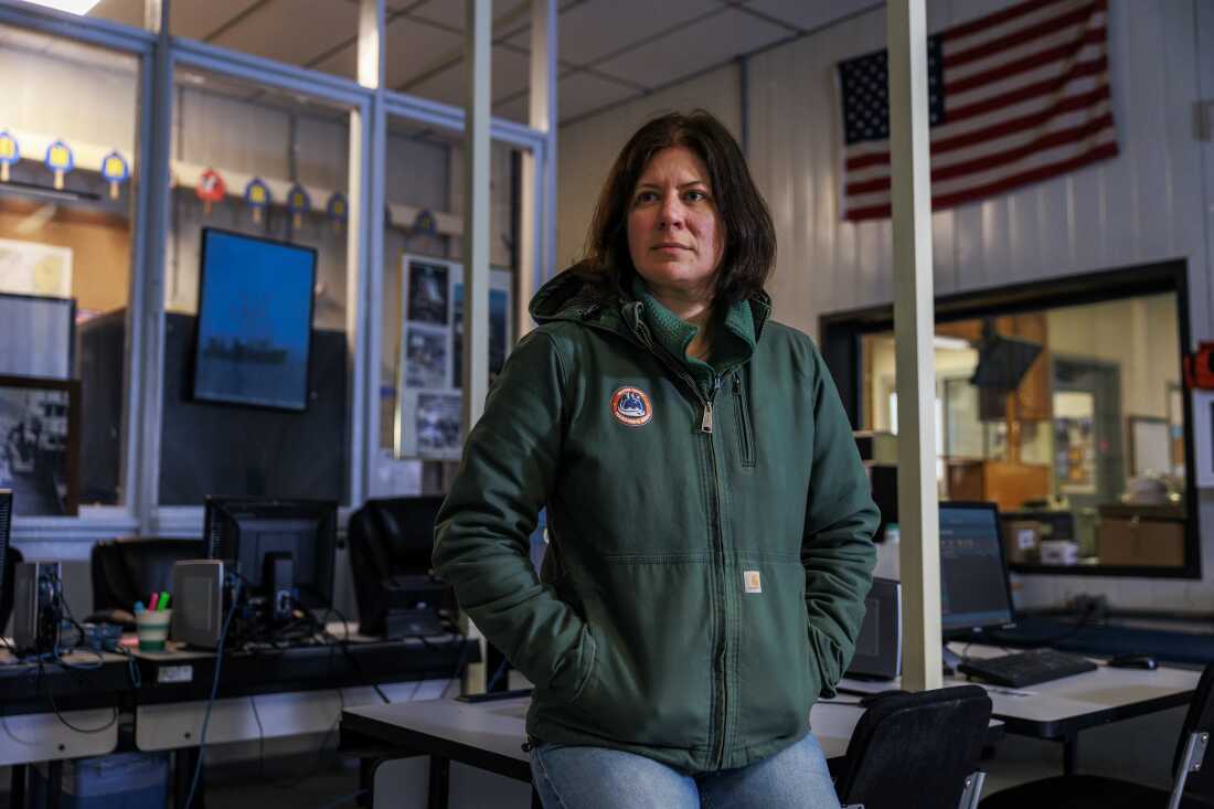 Wearing a green jacket and blue jeans, Mary Hudson is sitting on a desk inside a warehouse. Around her are other desks and workstations that have computer monitors on them.