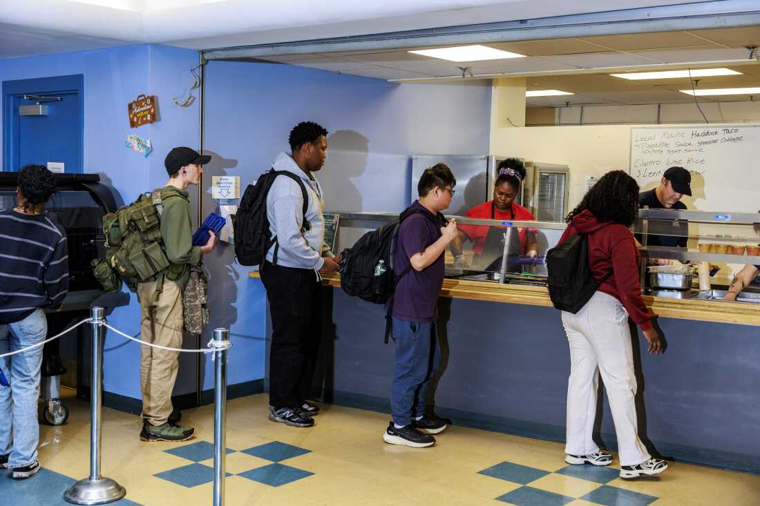 Five Westbrook High School students stand in a cafeteria line for lunch. Cafeteria workers stand on the other side of a clear divider, preparing food in metal containers.