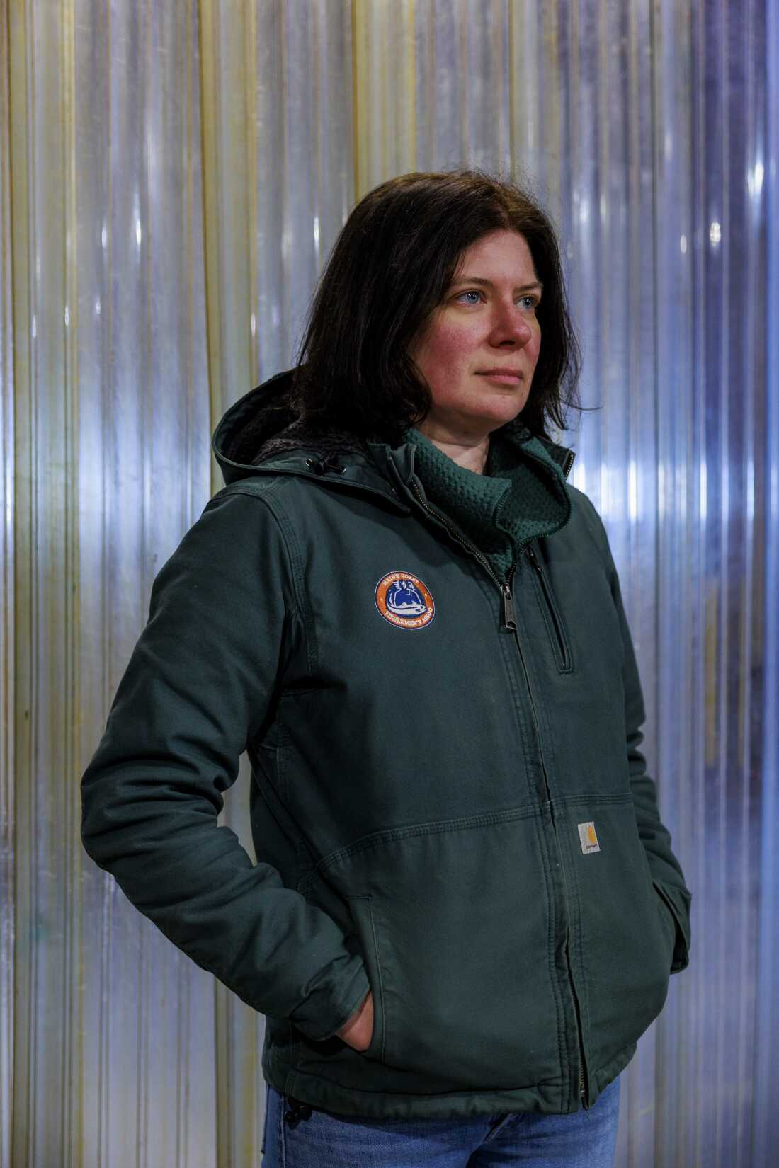 Mary Hudson stands in front of a plastic wall. She's wearing a green jacket and blue jeans.