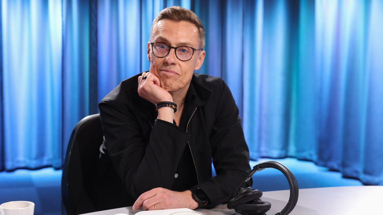 A man wearing glasses and a black zipper jacket sits with a half-smile in a radio studio with a headset on the table.