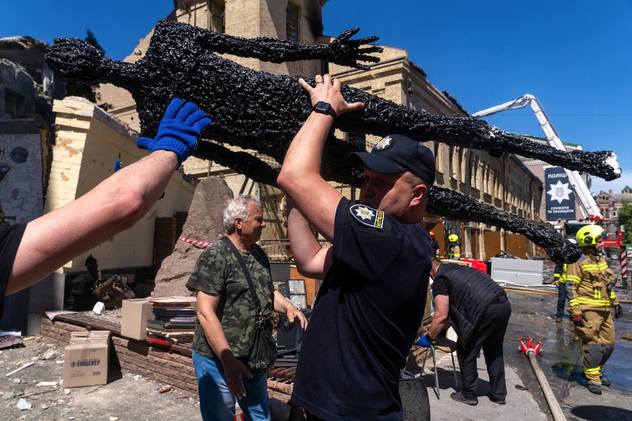 A man in a police uniform carries a black sculpture above his head as other people around him clean up at the scene.
