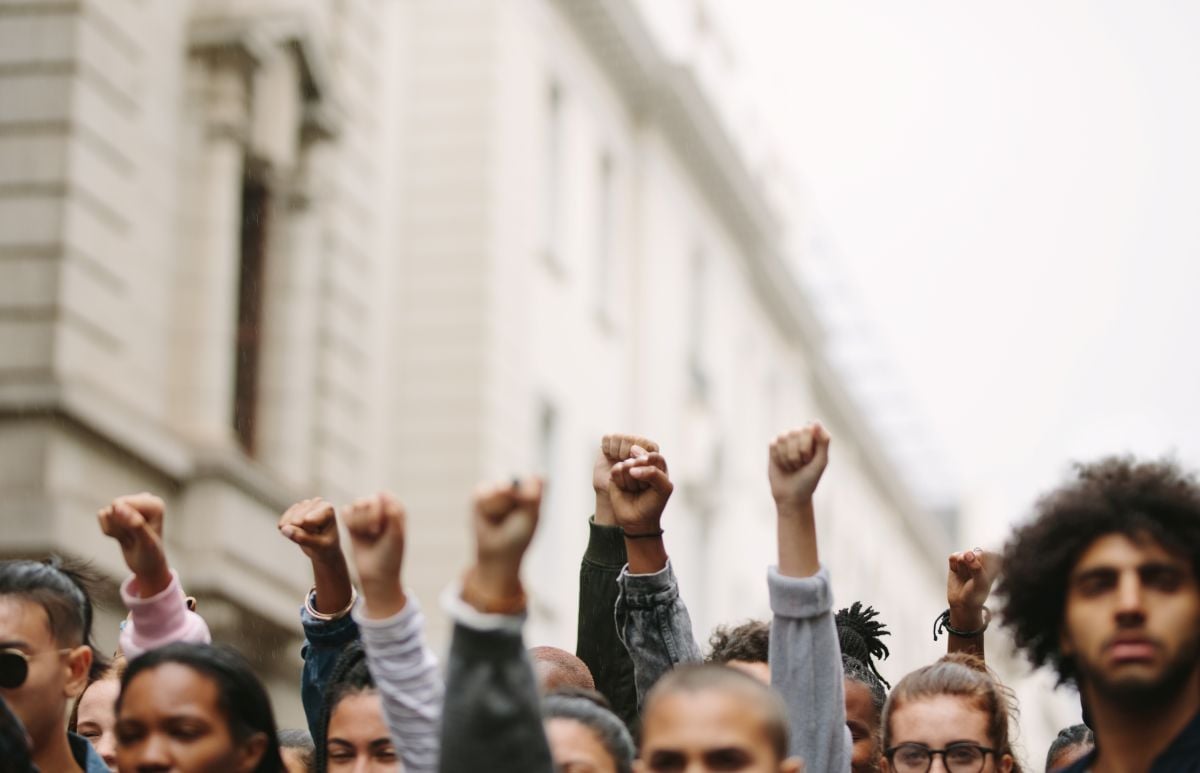 A group of people in front of a large white building hold up their fists in protest.