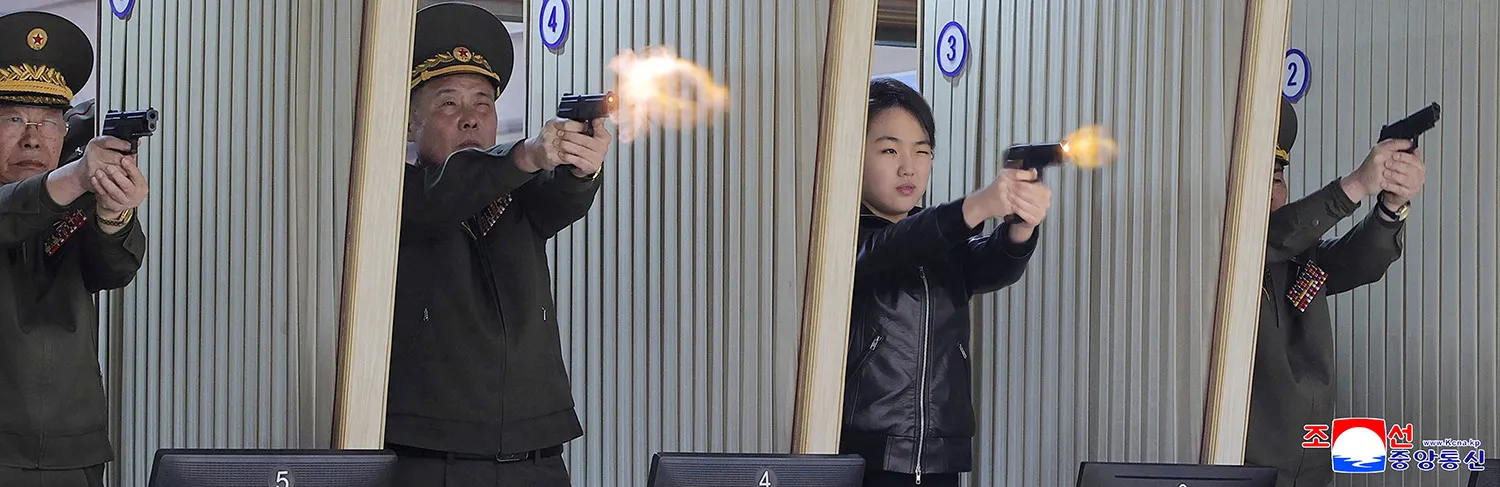 A girl in a black leather jacket and three men in military uniforms stand in separate firing booths at an indoor range. All four are holding handguns with both hands, arms extended, and aiming forward. Muzzle flashes and small puffs of smoke are visible at the ends of their barrels.