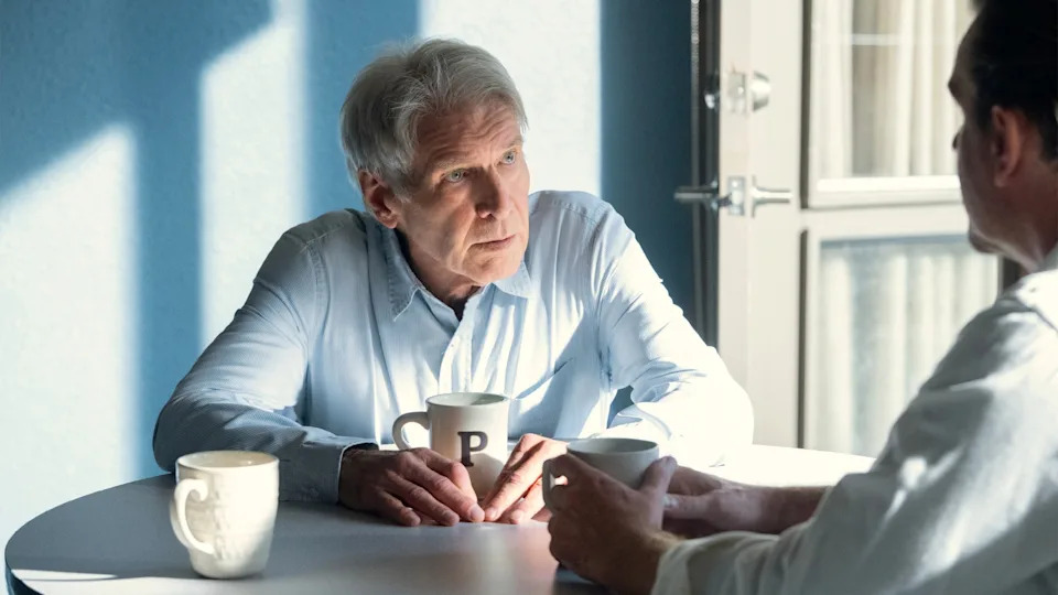 Two people sit at a table, each holding mugs. One listens intently, with light streaming through nearby windows