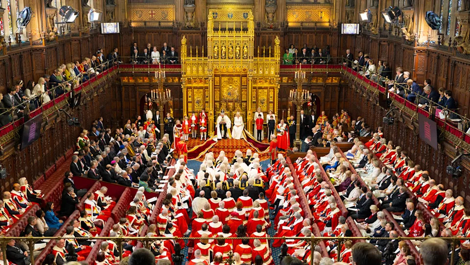 The House of Lords is a very grand looking wood panelled chamber with red benches. Many people are sitting wearing red robes in front of the King and Queen at the State Opening of Parliament.