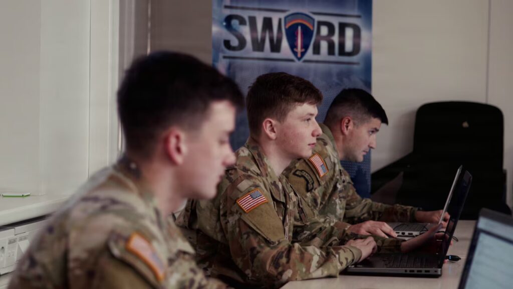 Three people in camouflage military uniforms sit indoors at a table, looking at computer screens.