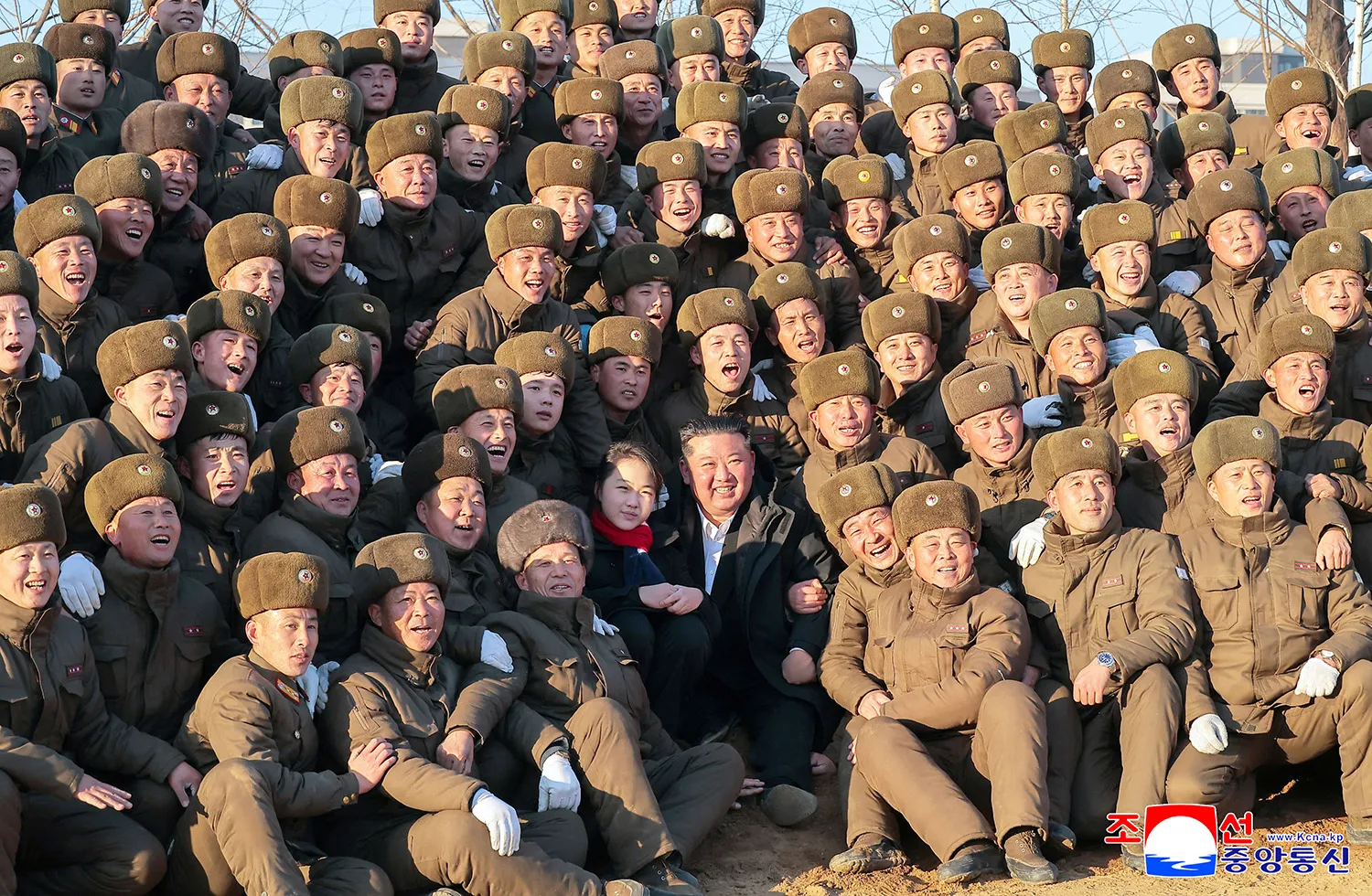 A man in a dark coat and a girl wearing a red scarf are surrounded by a large, dense crowd of men in brown military winter uniforms and fur-trimmed hats. Most of the men in the crowd are smiling and cheering, while the central pair looks toward the camera.