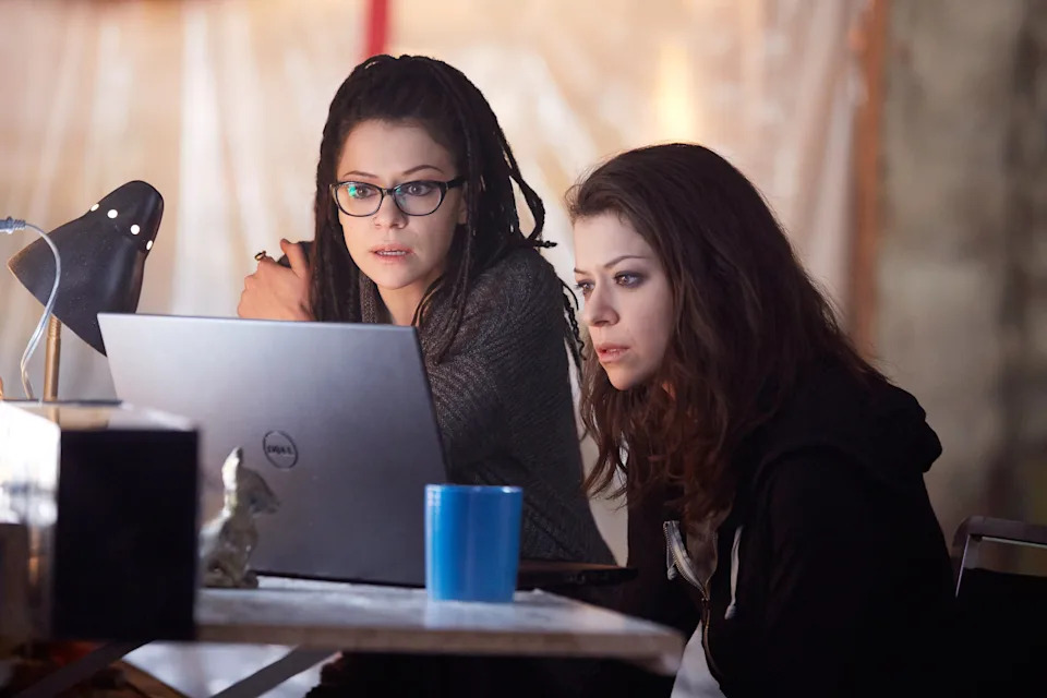 Two women in a dimly lit room focus intently on a laptop. One has glasses and a sweater, the other a hoodie
