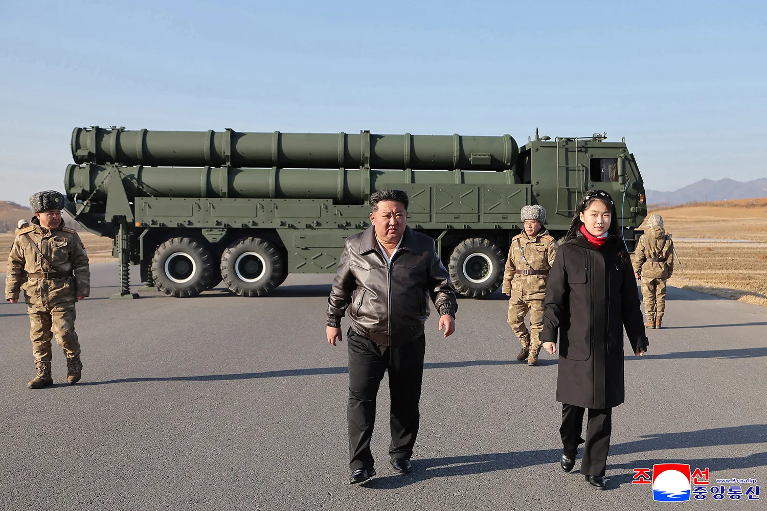 A man in a brown leather jacket and a girl in a long black coat walk across an asphalt field. In the background, a large olive-green military truck carries several long, cylindrical canisters. Two men in camouflage winter gear walk behind them in an open, hilly landscape under a clear sky.