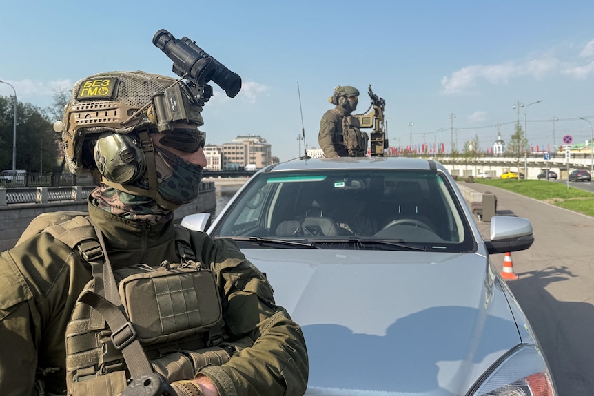 A soldier wearing protective gear and a helmet in front of a truck with another soldier armed with a machine gun.
