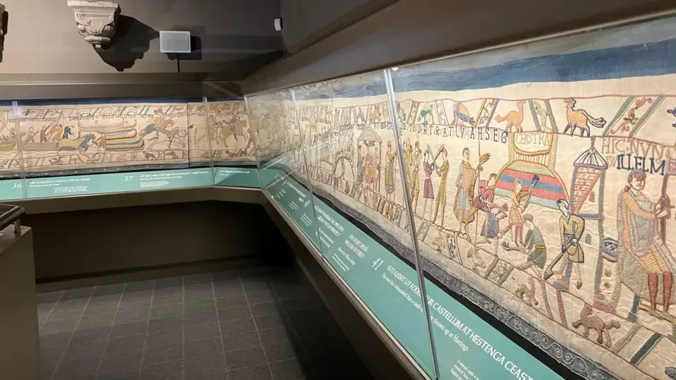 The Bayeux Tapestry depicting the Norman conquest in 1066 is pictured behind a glass case along a grey wall.