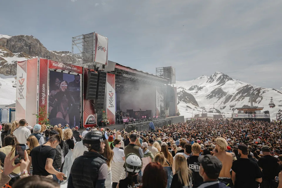 Outdoor concert with a large crowd in a snowy mountain setting. A performer is on stage with visuals displayed on large screens