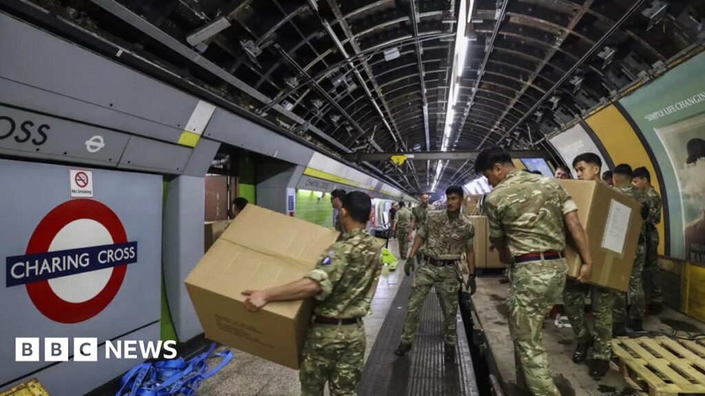 Soldiers in combat gear move boxes off pallets on a disused Tube platform.