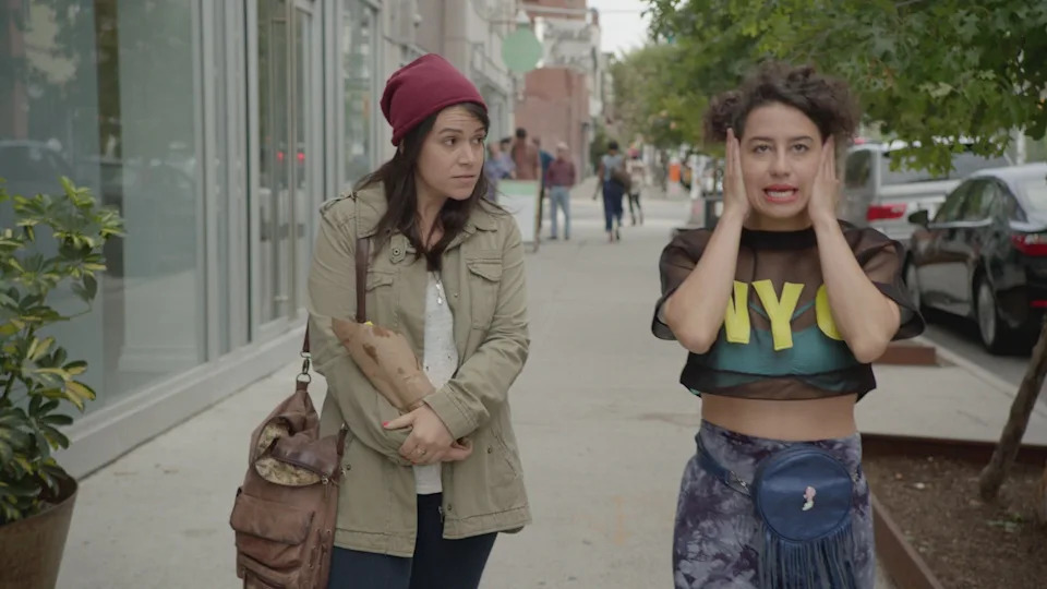 Two women walking on a sidewalk, one wearing a beanie and holding a bag, the other in a crop top with "NYC" text, looking surprised