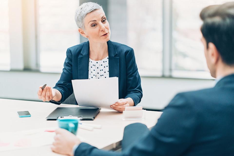 Two investors sit at a conference table while one holds some papers and explains something to the other.