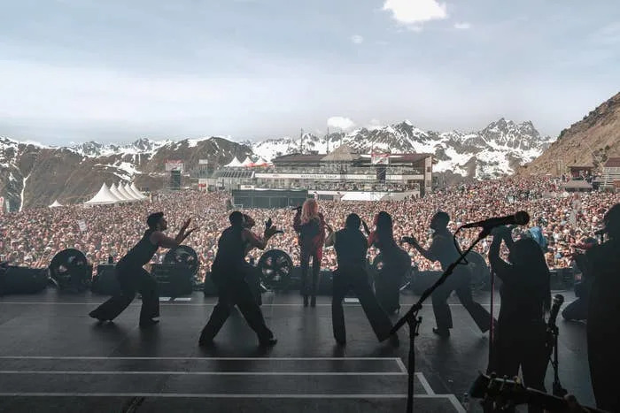 Singer performing to a large outdoor crowd in a mountainous setting, with backup dancers and snow-capped peaks in the background