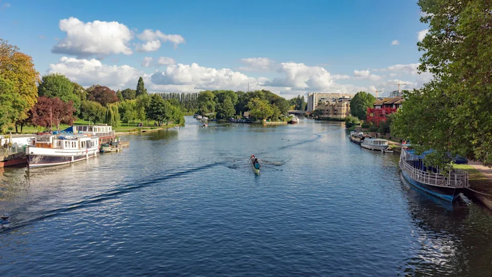 Rowers navigate the River Thames in Reading. The large Christchurch Meadows park is to the left and Reading town centre is to the right. People can be seen in the distance enjoying the park on a sunny autumnal day.