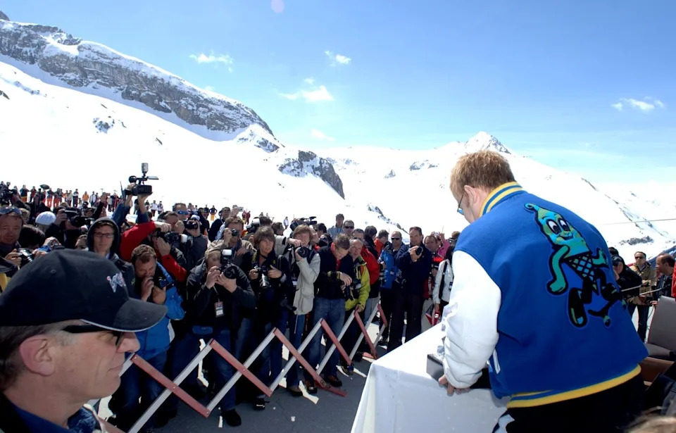 Person in a jacket featuring a cartoon character, standing at a podium, addressing a crowd of photographers and onlookers in a snowy mountain setting