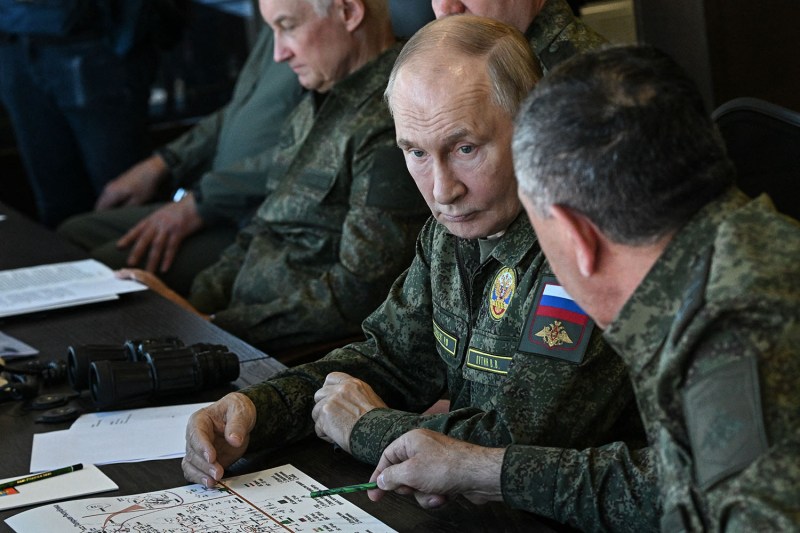 A close-up of several individuals in camouflage military uniforms seated at a table. One man in the center looks intensely toward another person whose back is partially to the camera. On the table, several papers with tactical diagrams and a pair of binoculars are visible.
