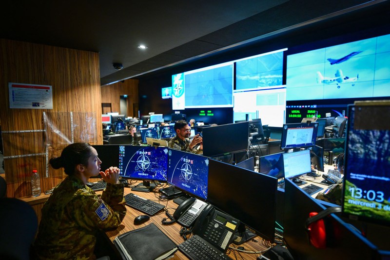 The interior of a dimly lit, modern operations center filled with multiple computer monitors and large wall displays. Personnel in military uniforms sit at workstations, and one woman in the foreground gazes toward the screens. The displays show various digital maps, aircraft footage, and organizational logos.