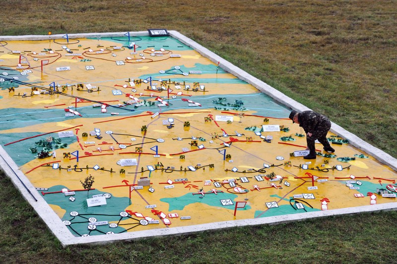 A large, colorful relief map lies on a grassy field, enclosed by a white concrete frame. The map features sections of yellow, green, and blue, marked with various tactical symbols, small flags, and miniature military models. A person in camouflage uniform leans over the map, placing a small white marker near a cluster of symbols.