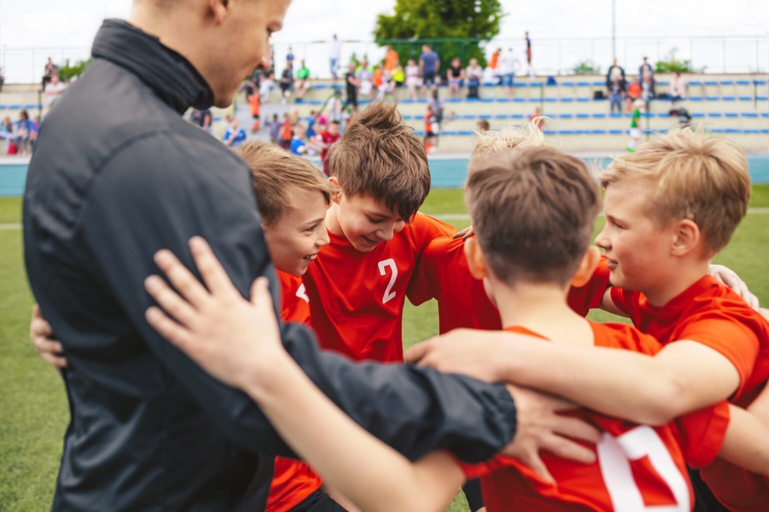 Sports coaches can improve health for athletes with disabilities Happy boys in sports team huddling with young coach. Kids standing in a circle on grass sports pitch. Children sports team