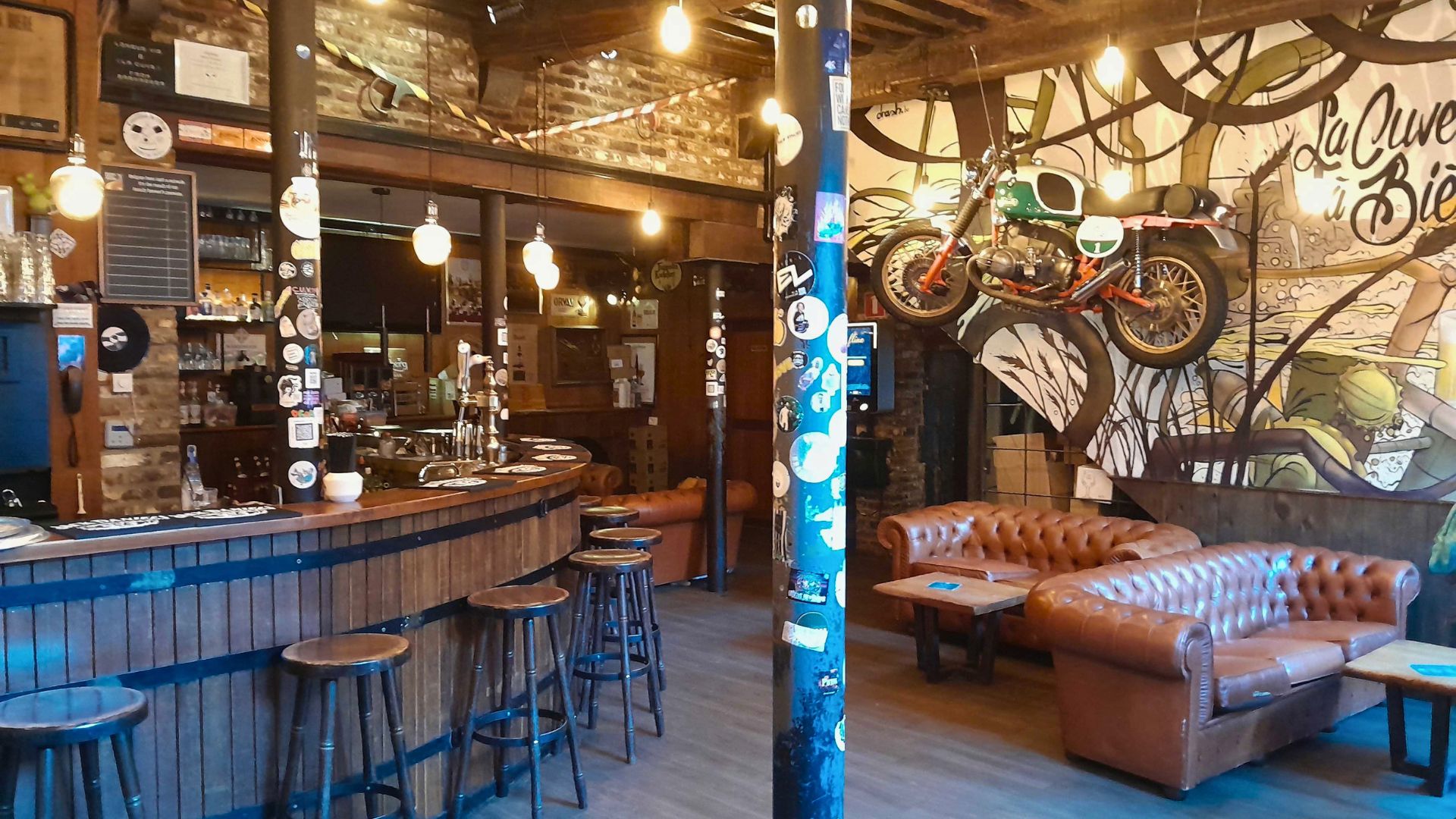 Interior of La Cuve &agrave; bi&egrave;re. Rustic bar interior with a wooden curved bar, high stools, leather sofas, and artistic graffiti. A vintage motorcycle hangs on exposed brick, lit by warm bulbs