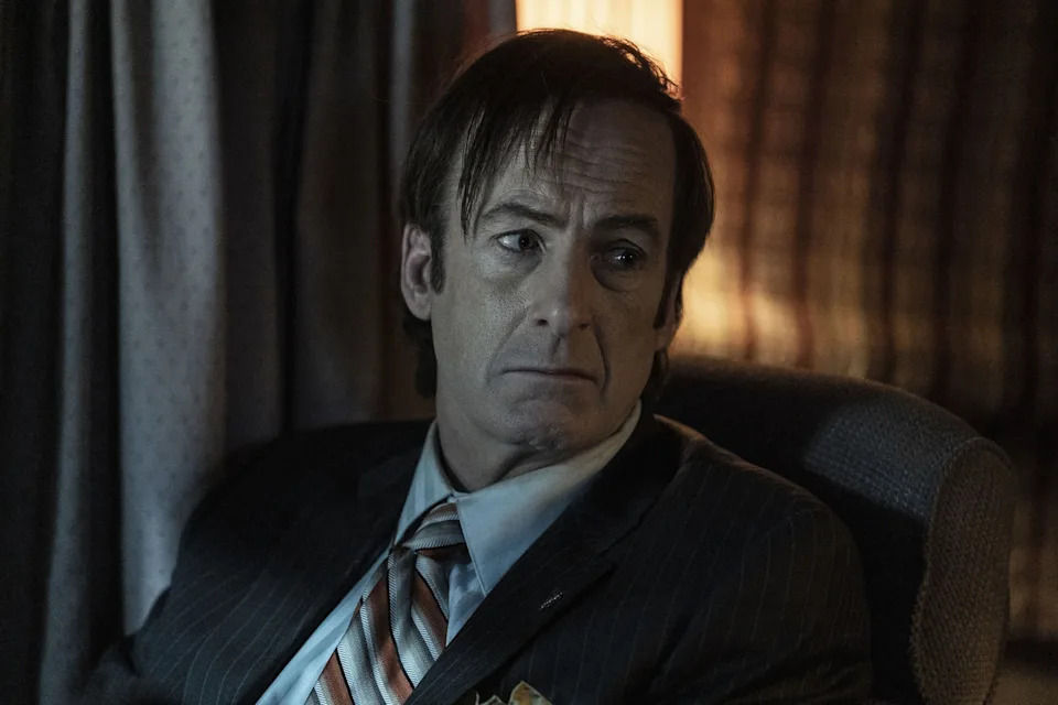 A man in a pinstripe suit and striped tie sits pensively in a dimly lit room, likely depicting a scene from a TV drama