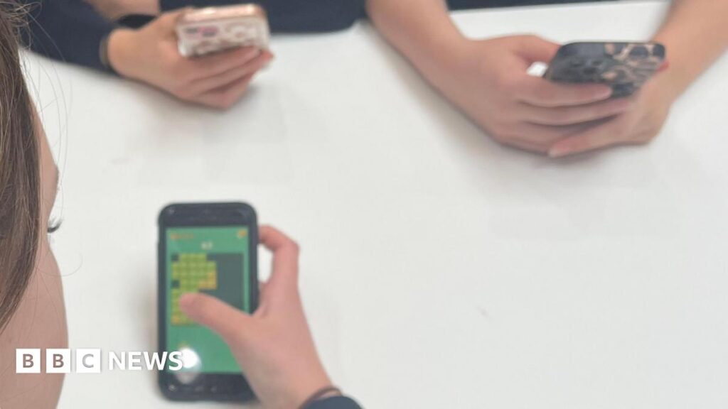 Phone-free school event to bring in guest speakers A picture of schoolchildren on their mobile phones, sat surrounding a white table.