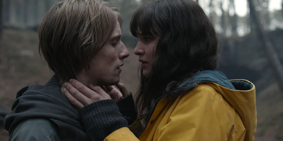 Two people stand close in a forest, gazing into each other's eyes. One wears a yellow raincoat. Emotional moment likely from a film or TV show