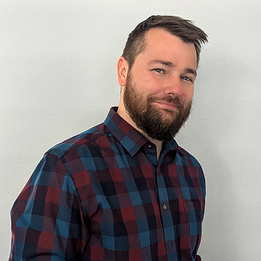 Ben Lyons, wearing a plaid shirt leans sideways and smiles towards the camera